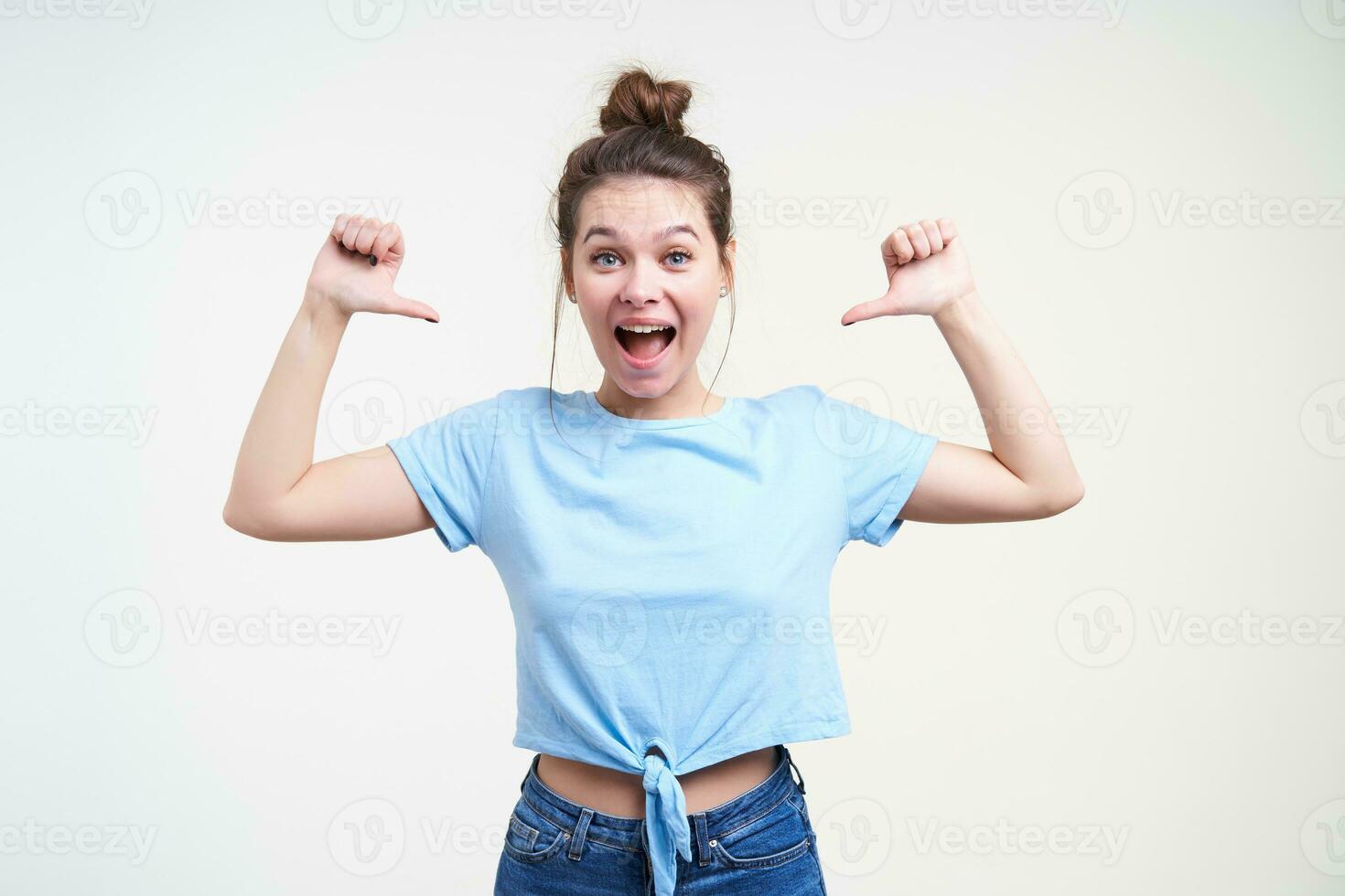 Agitated young pretty dark haired female with natural makeup keeping her mouth wide opened while showing happily on herself, standing over white background photo