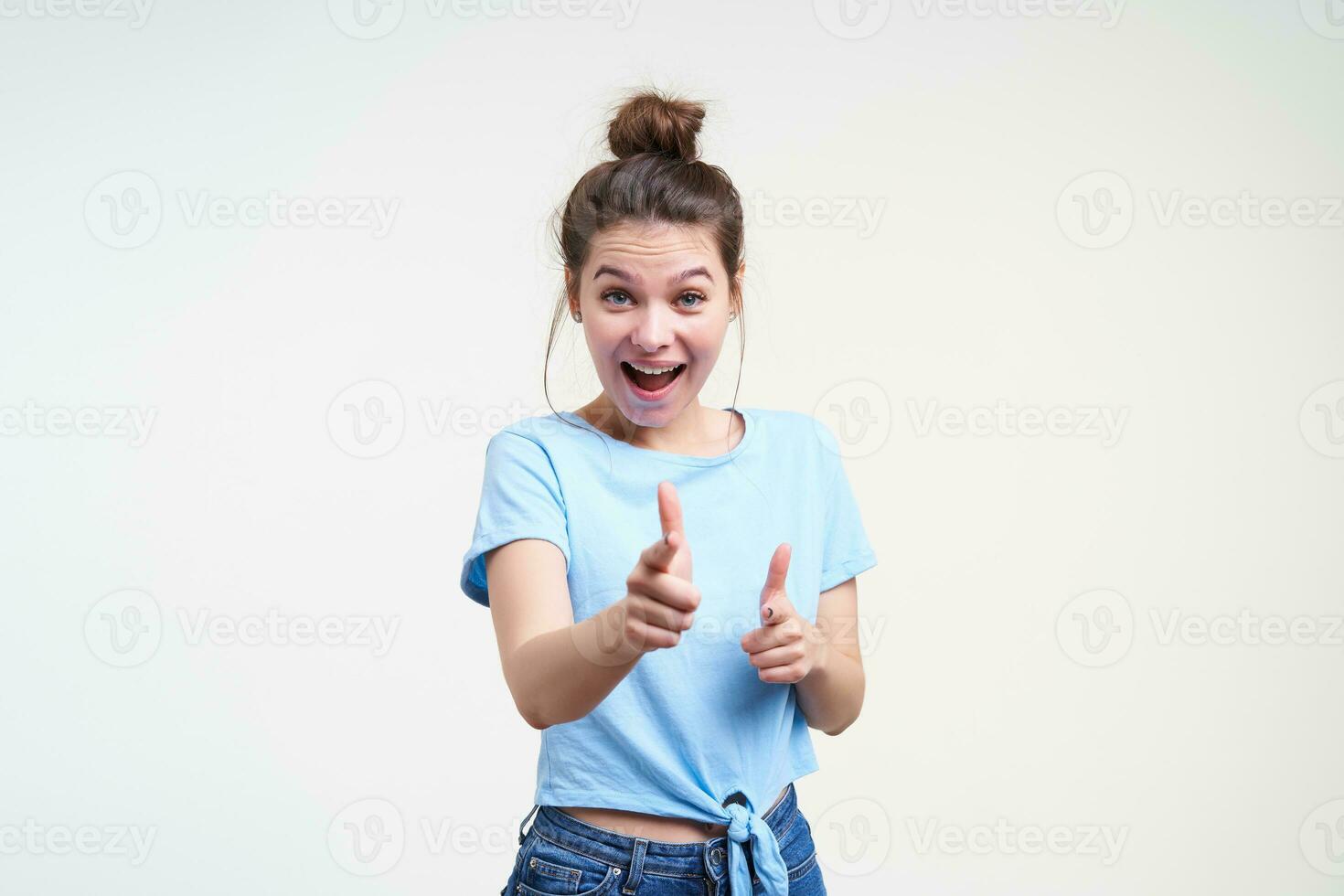 alegre joven bonito morena mujer con casual peinado acuerdo manos elevado mientras señalando alegremente a cámara, en pie terminado blanco antecedentes en azul camiseta foto