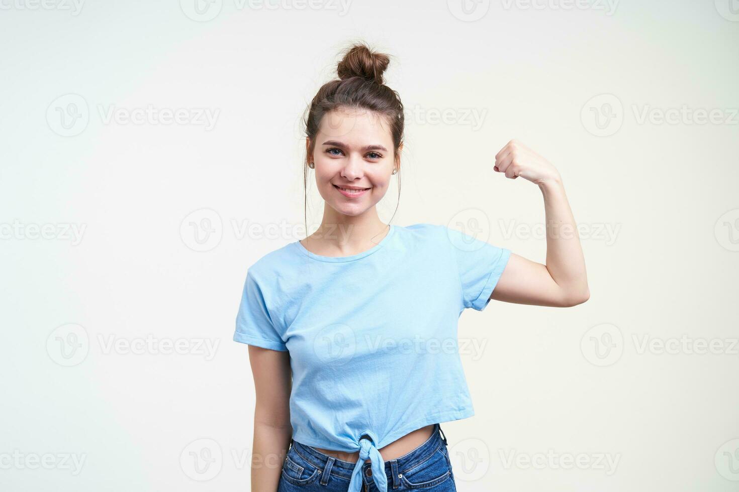 Cheerful young lovely brunette lady with casual hairstyle smiling gladly while showing her strong biceps, standing over white background in blue t-shirt photo