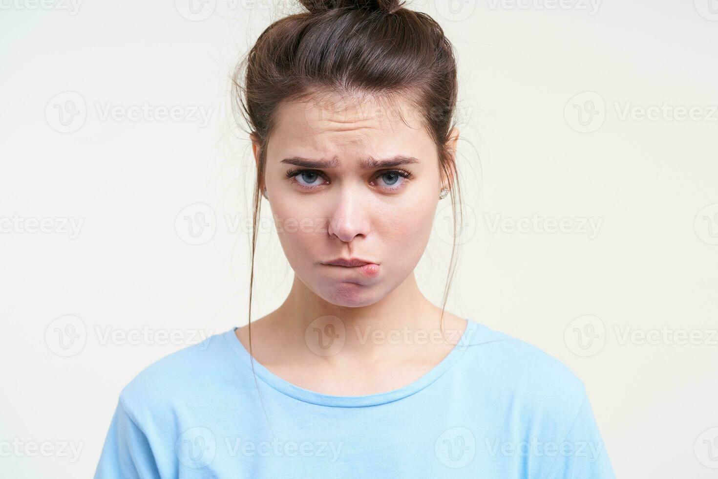 Close-up of young beautiful unhappy brown haired lady with blue eyes frowning her eyebrows and biting underlip while looking sadly at camera, posing over white background photo