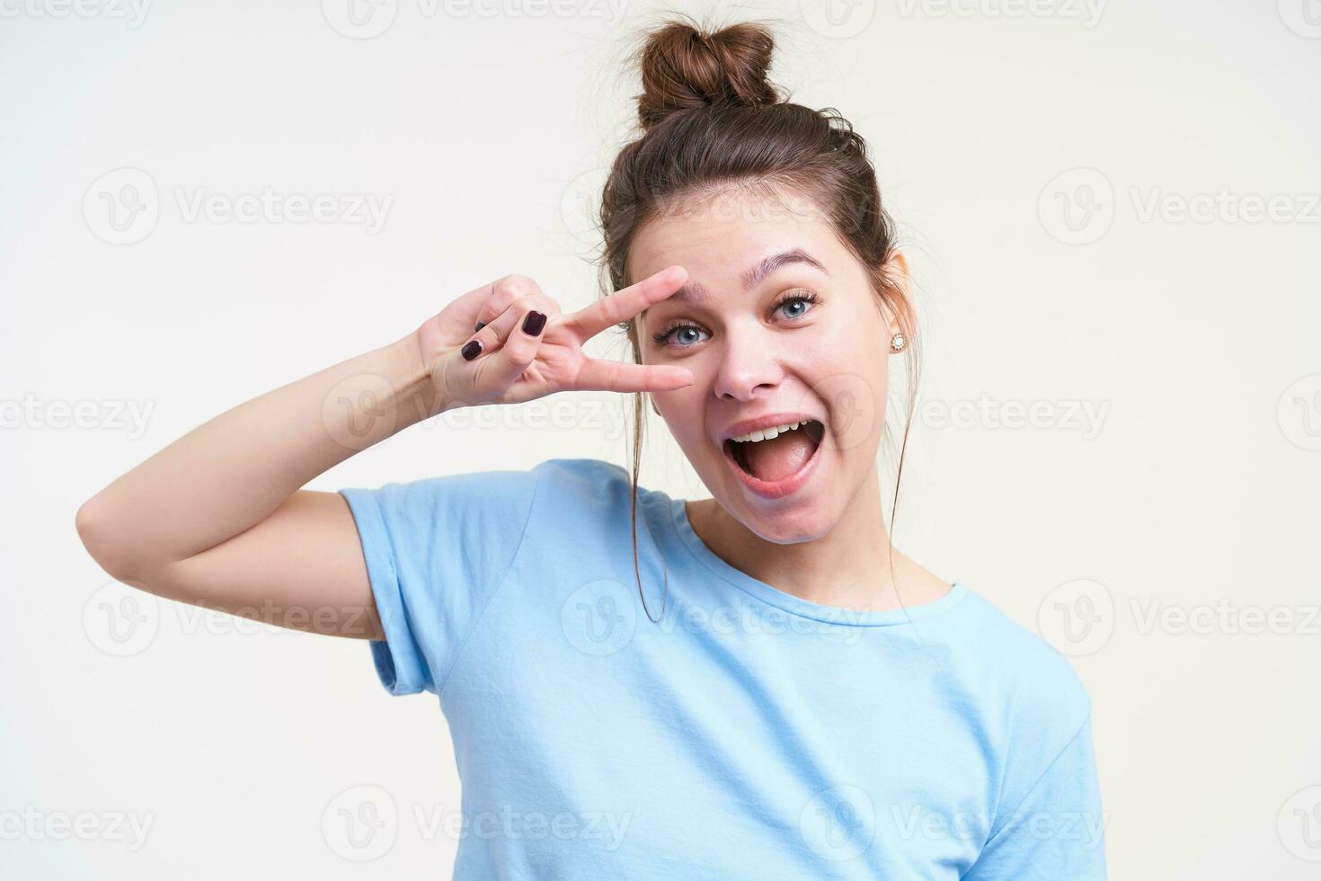 Excited young lovely brown haired woman raising hand with victory gesture and looking at camera with wide mouth opened, standing over white background photo