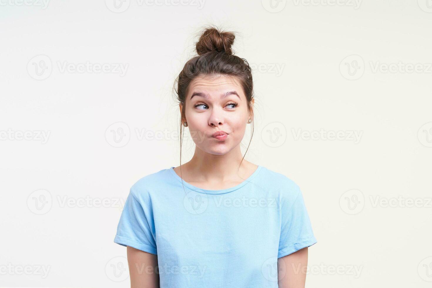 Puzzled young lovely brunette lady with natural makeup raising her eyebrows while looking wonderingly aside and pouting lips, posing over white background photo