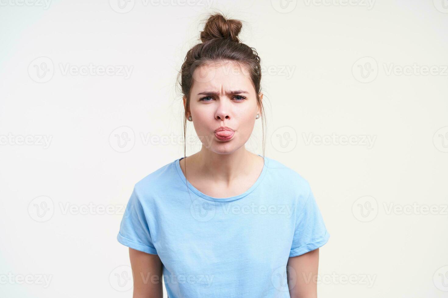 Studio photo of young gloomy dark haired woman with bun hairstyle frowning her eyebrows and showing tongue while standing over white background with hands down