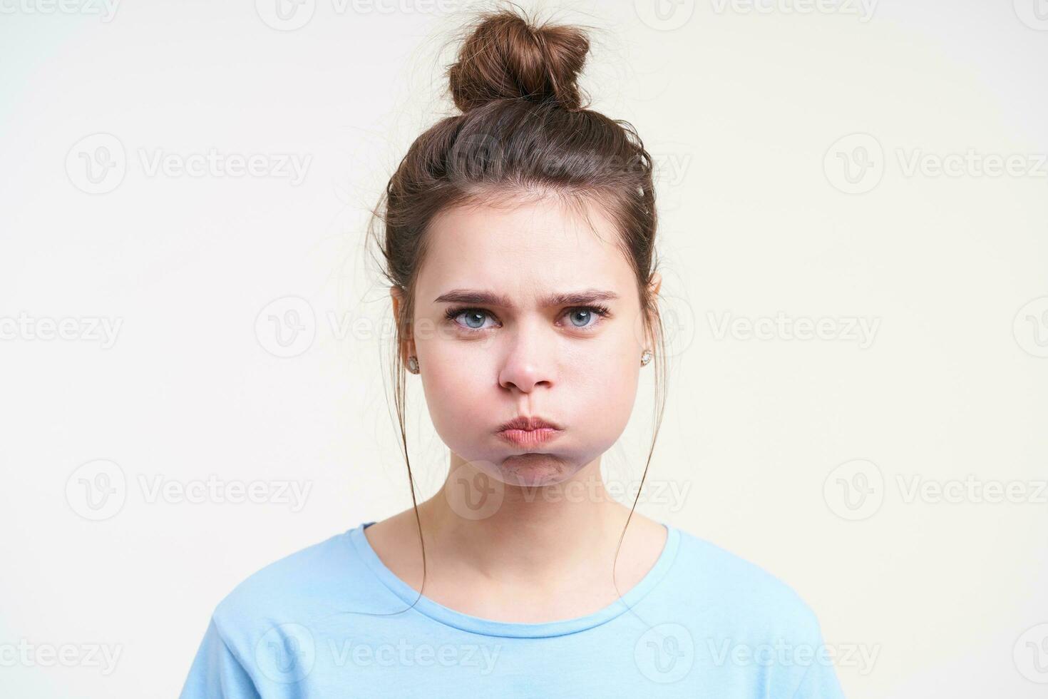 Portrait of young blue-eyed pretty brunette female taking air into her mouth and puffing out cheeks while looking at camera, standing over white background photo