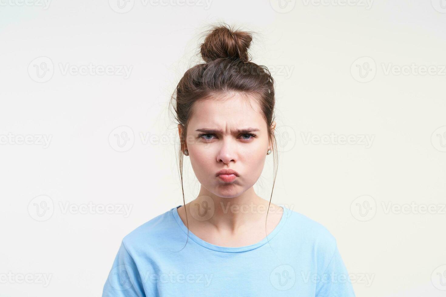 Severe young attractive brown haired lady squinting her eyes and pouting lips while looking crossly at camera, posing over white background in casual clothes photo