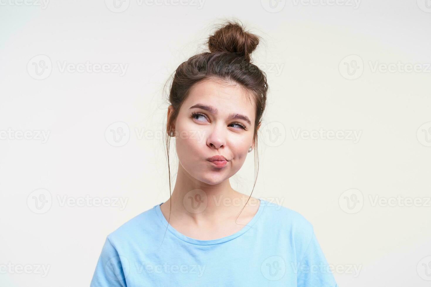 Indoor shot of young pensive brown haired lady with natural makeup twisting her mouth while looking thoughtfully aside, isolated over white background photo