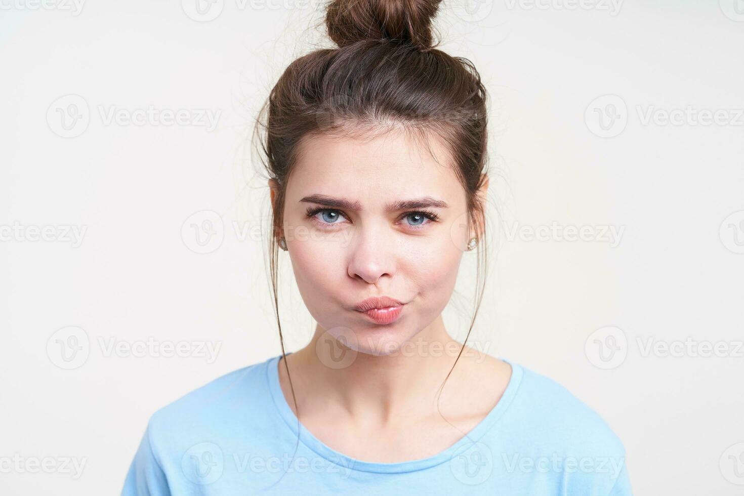 Close-up of puzzled young pretty brown haired woman with bun hairstyle twisting her mouth while looking at camera, standing over white background in casual wear photo