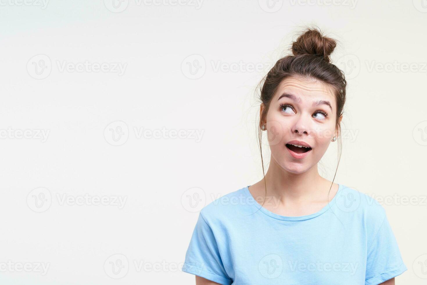 Puzzled young attractive brown haired lady with casual hairstyle looking wonderingly upwards with opened mouth while standing against white background photo