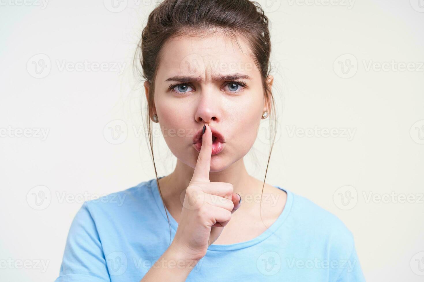 Portrait of severe young pretty brunette female frowning her eyebrows and raising hand with hush gesture to her mouth, standing against white background photo