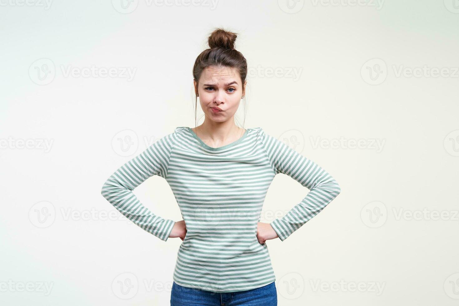 Puzzled young blue-eyed pretty brown haired lady twisting her mouth while looking at camera and holding hands on her waist while posing over white background photo