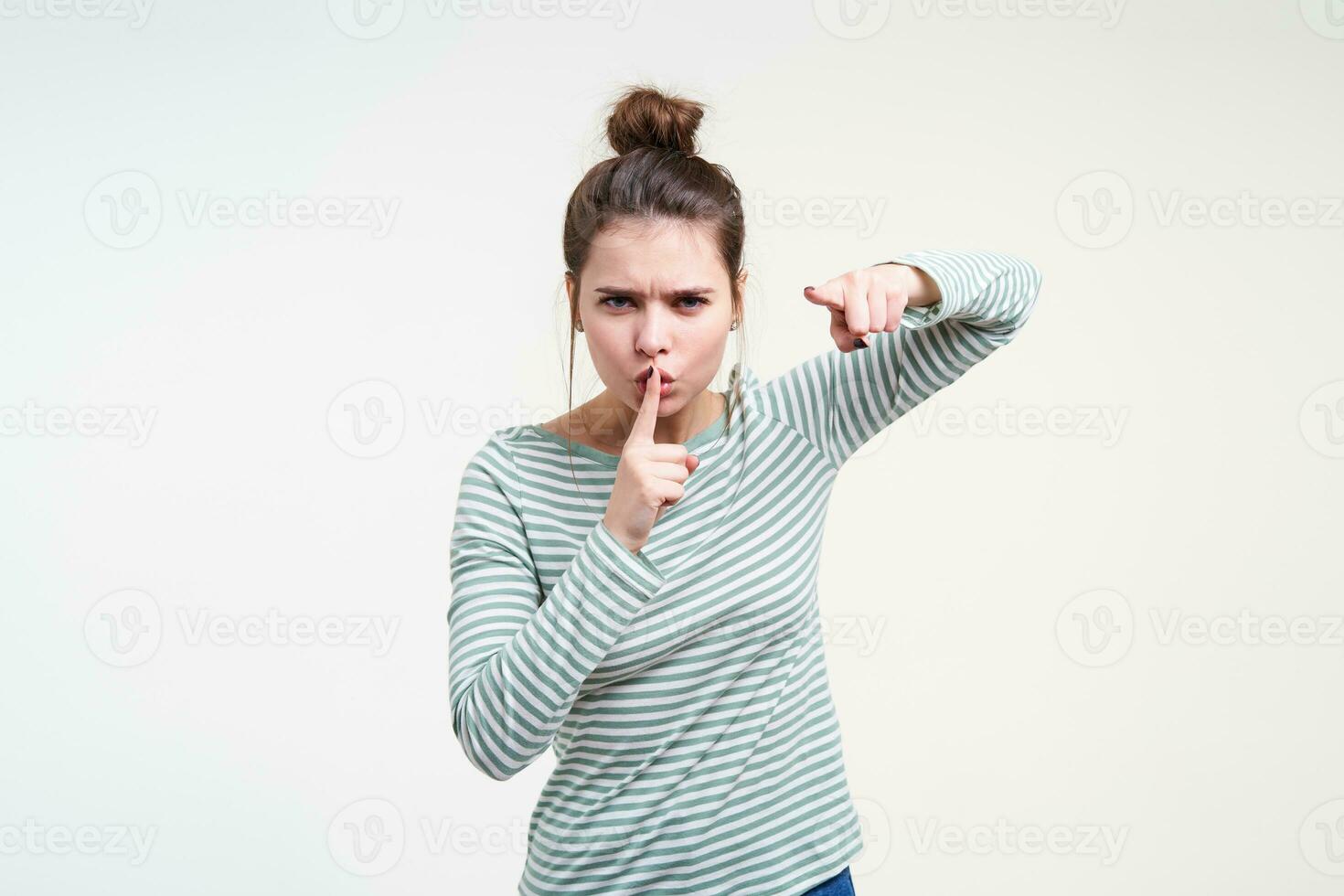 Severe young pretty brunette woman with bun hairstyle pointing angrily at camera and raising hand in hush gesture while standing over white background photo