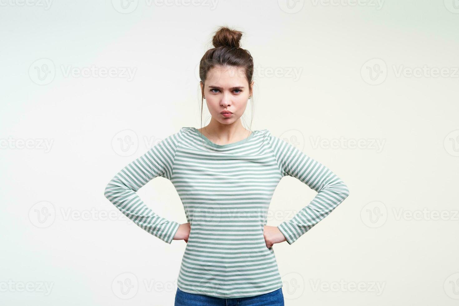 Serious young attractive brunette woman with casual hairstyle looking crossly at camera and keeping hands on her waist while posing over white background photo