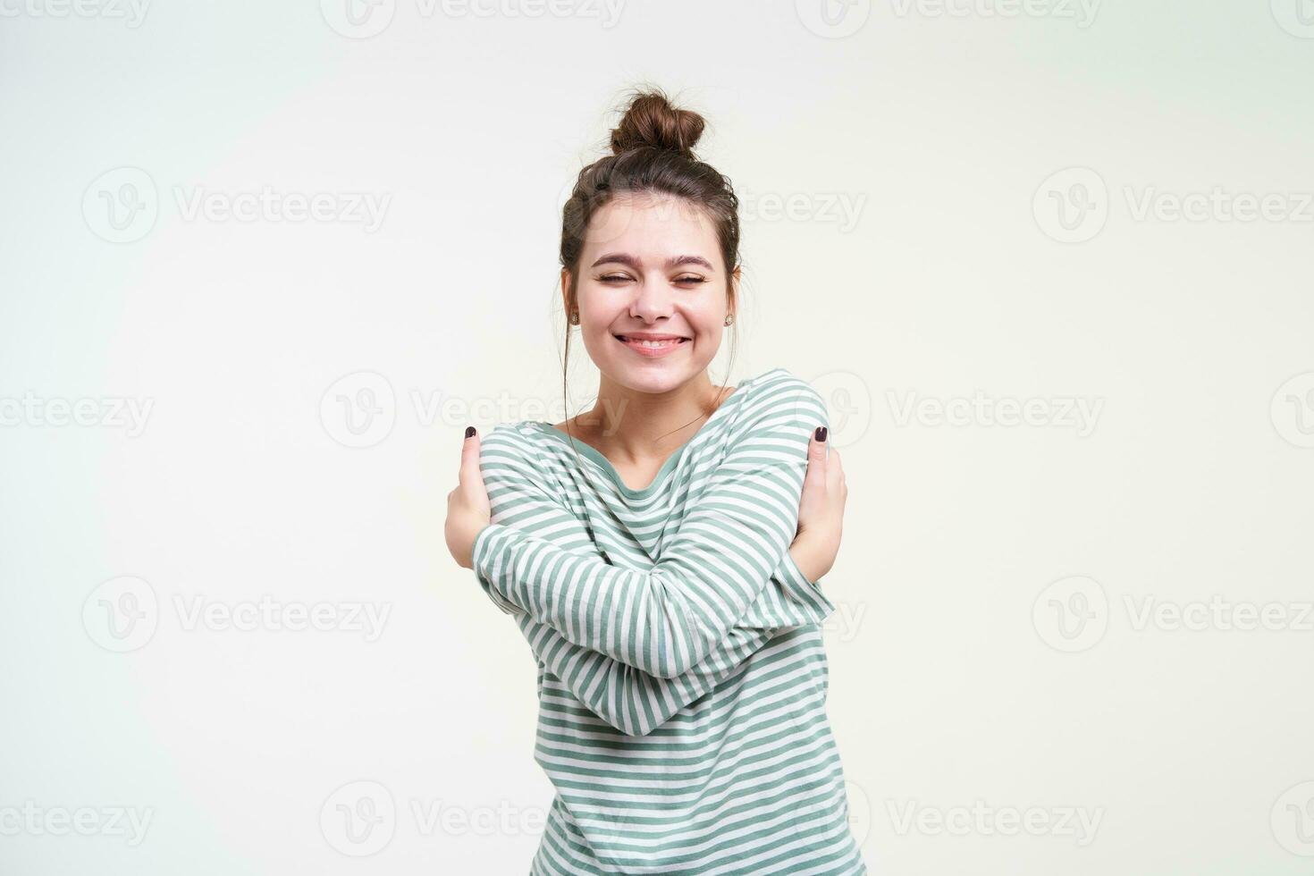 Pleased young lovely brown haired lady keeping her eyes closed while hugging herself and smiling pleasantly, standing over white background in striped blouse photo