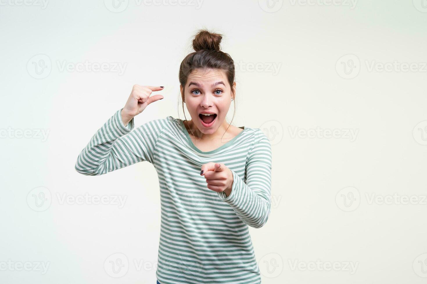 Bemused young blue-eyed pretty brunette female pointing amazedly at camera with forefinger and showing small size with other hand, isolated over white background photo