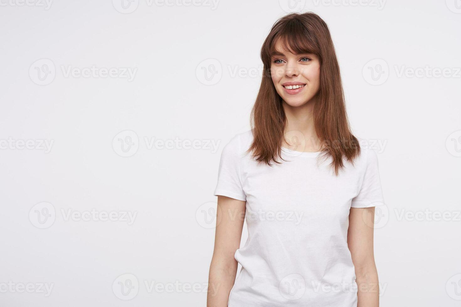 Positive young attractive woman with natural makeup looking cheerfully aside with pleasant smile, keeping hands along body while standing over white backgrounnd photo