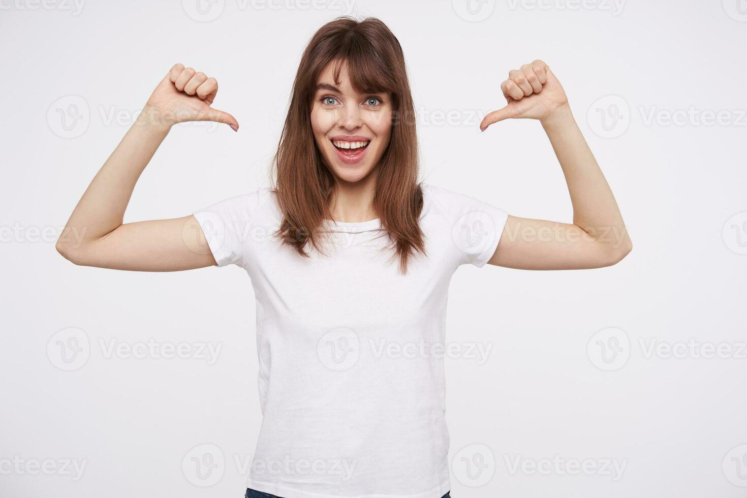 Cheerful blue-eyed lovely young brown haired lady smiling widely while showing on herself with raised hands, wearing basic white t-shirt while standing over white backgrounnd photo