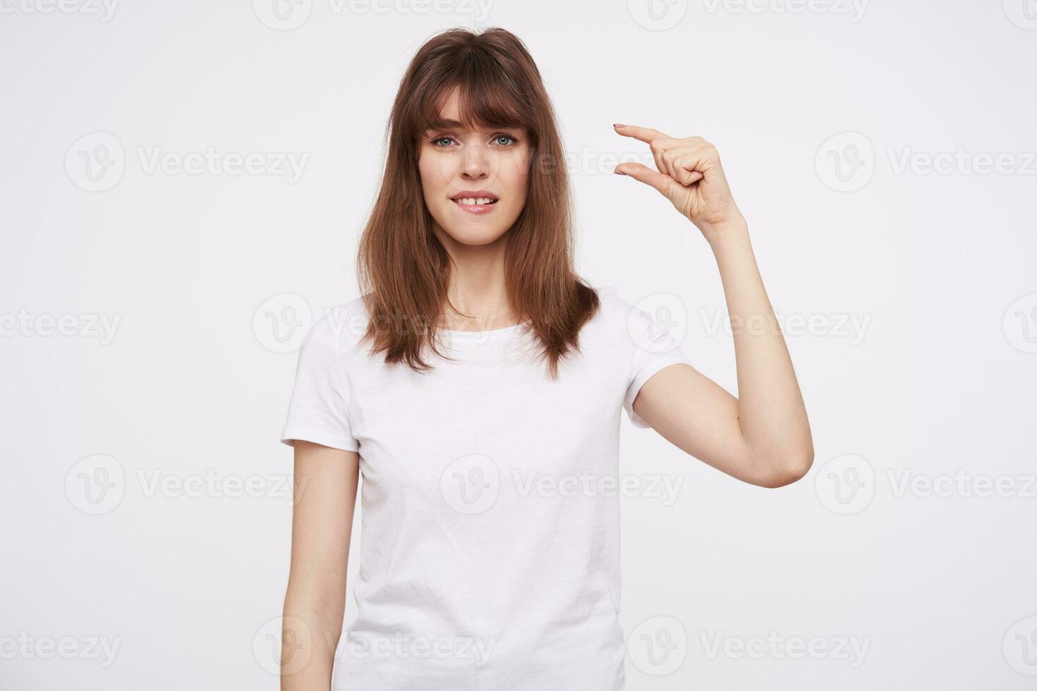 Portrait of young disappointed attractive brunette female with casual hairstyle showing small size with her fingers and biting underlip while looking to camera, isolated over white backgrounnd photo