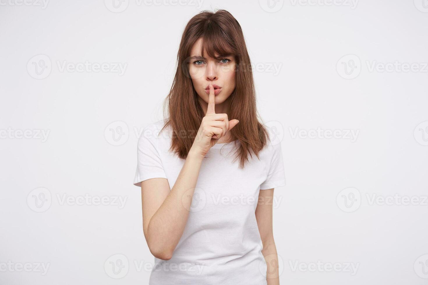Serious young pretty brunette woman with casual hairstyle frowning her eyebrows while asking to keep silence, raising hand in hush gesture while posing over white backgrounnd photo