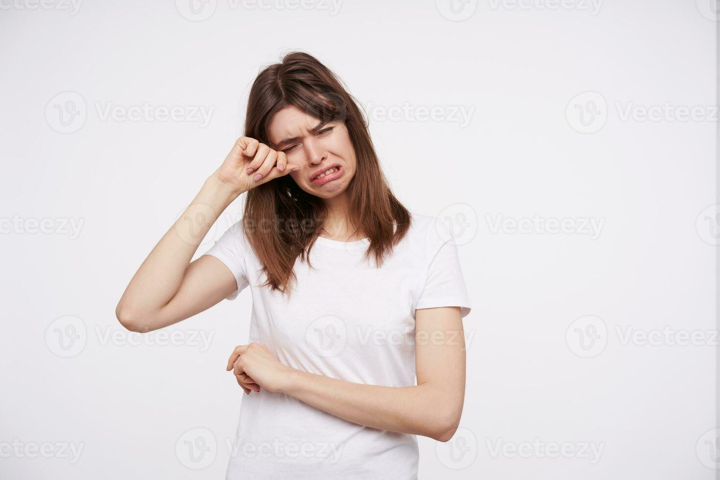 Unhappy young dark haired female dressed in white basic t-shirt keeping her eyes closed while crying and wiping away tears from her face, isolated over white background photo