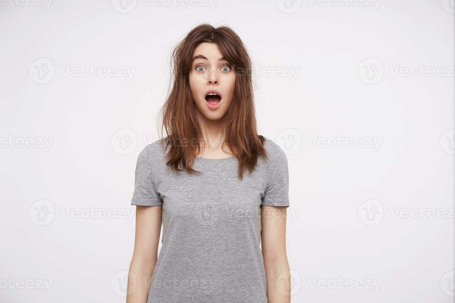 Shocked young blue-eyed pretty lady with natural makeup raising surprisedly eyebrows and keeping mouth wide opened while standing over white background photo