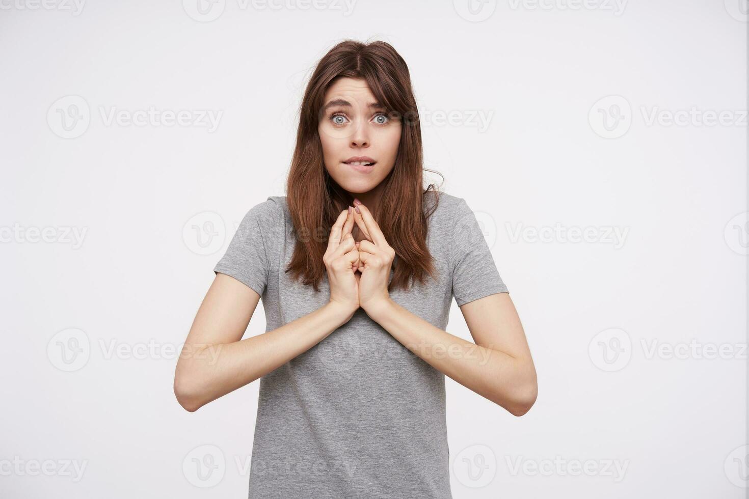 Excited young green-eyed brunette female biting worringly lips and keeping fingers crossed while looking at camera, isolated over white background in grey t-shirt photo