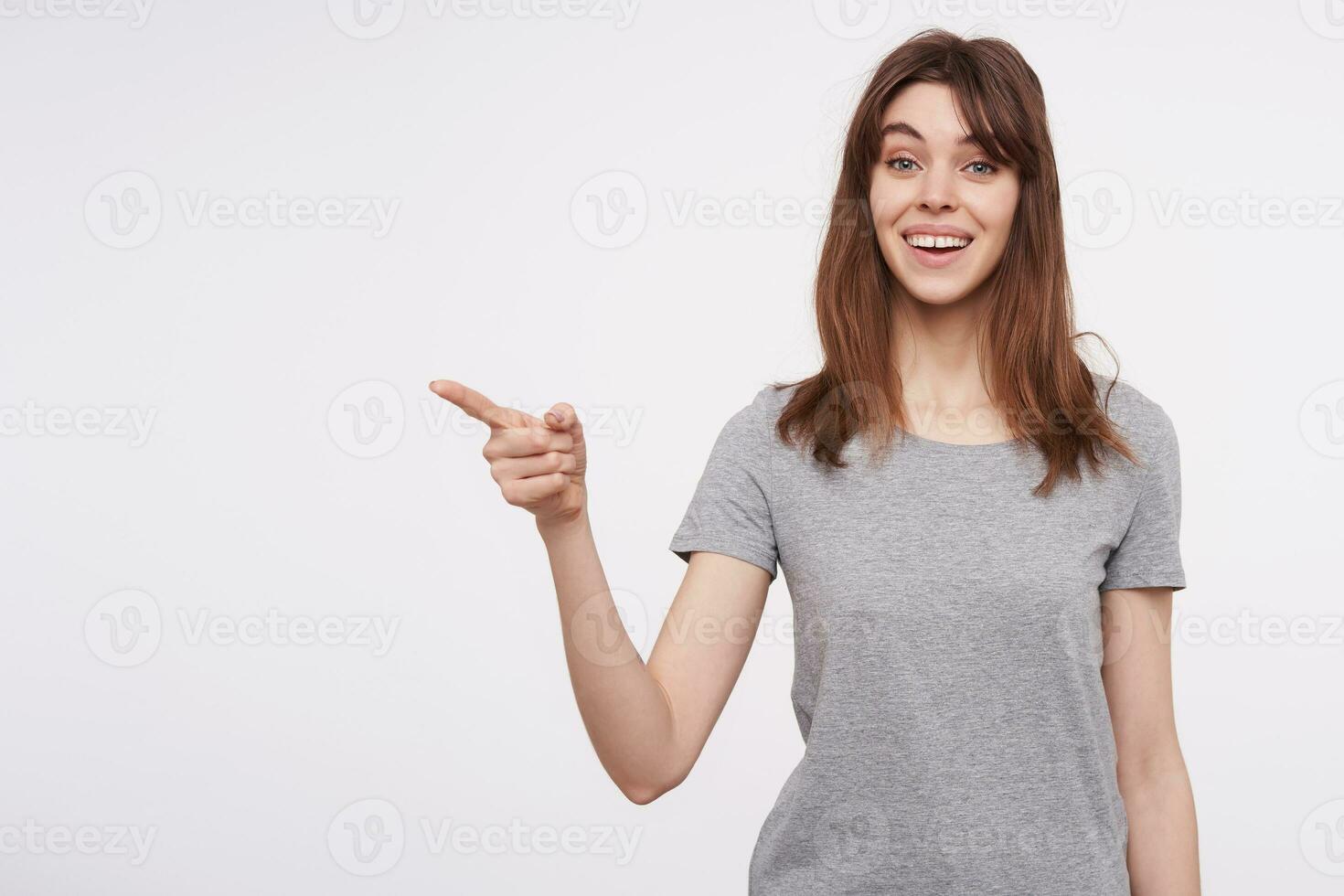 Indoor photo of young cheerful brunette woman with natural makeup smiling widely while pointing aside with forefinger, isolated against white background