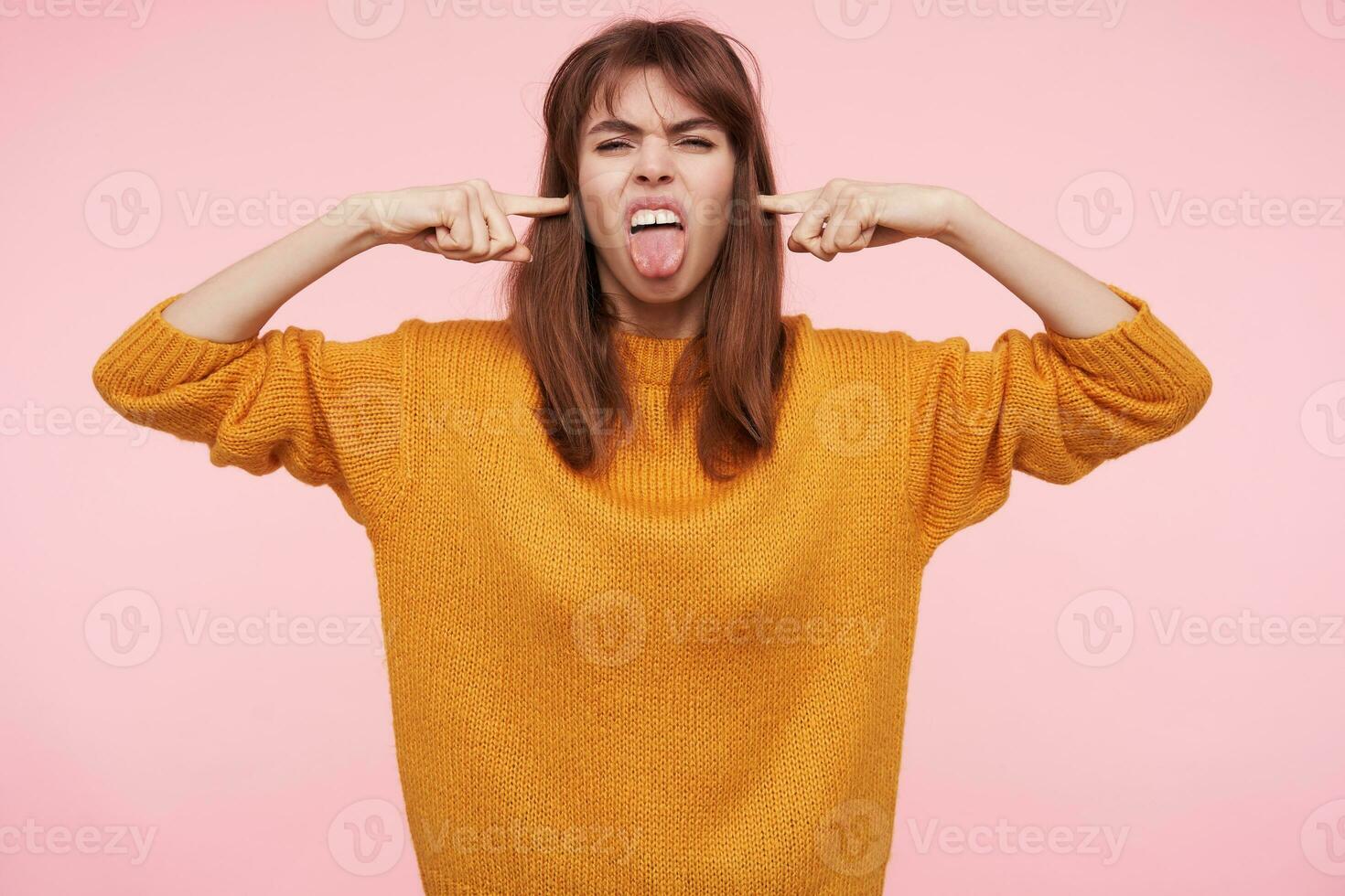 Horizontal photo of young dark haired woman with casual hairstyle grimacing her face and sticking out tongue while holding forefingers in her ears, isolated over pink background