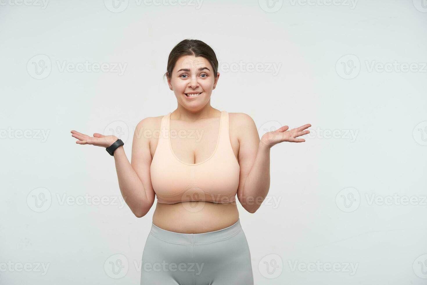 Studio photo of young oversized female in sporty clothes keeping her palms raised and looking perplexedly at camera, standing against white background