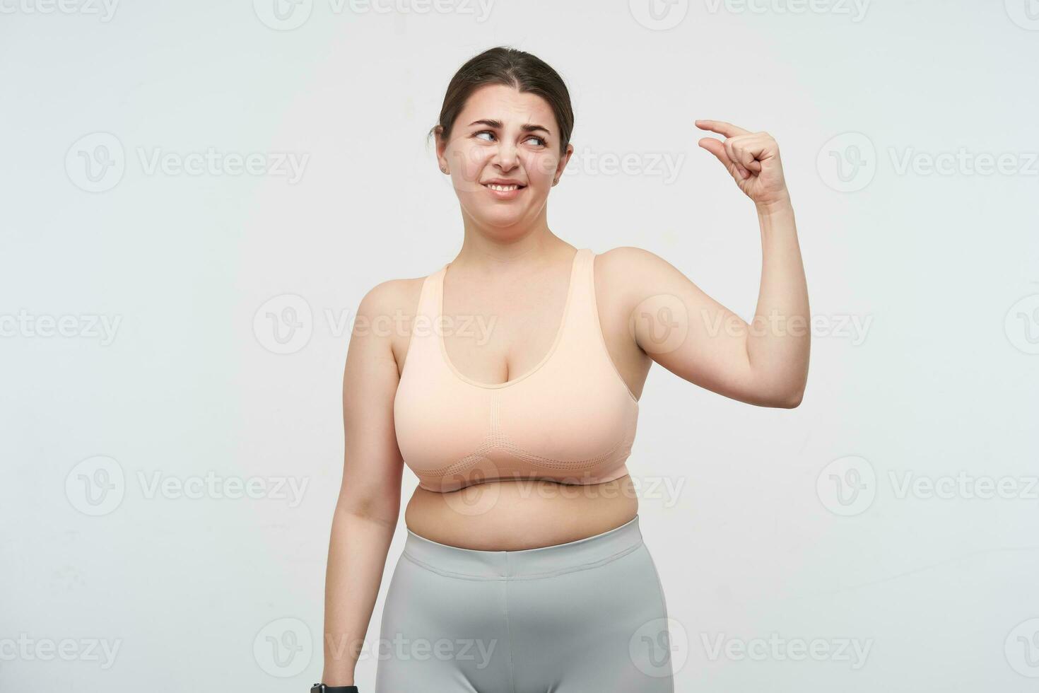 Puzzled young dark haired plump woman with ponytail hairstyle showing small size with raised hand and frowning her faced, standing against white background photo
