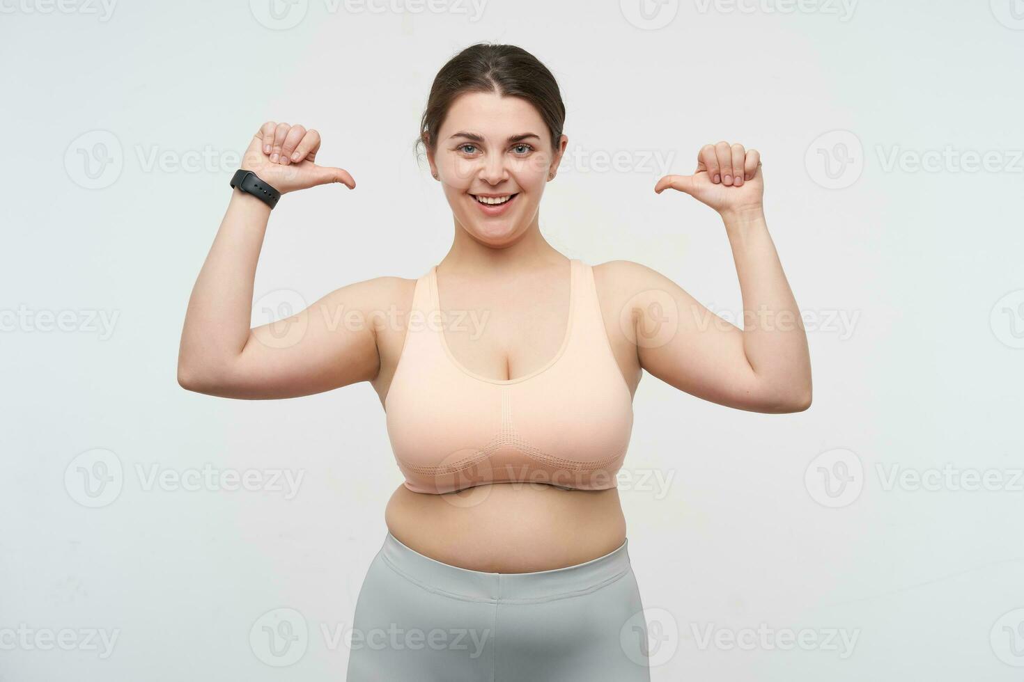 Studio shot of young happy cute dark haired fatty lady with ponytail hairstyle thumbing on herself while looking cheerfully at camera, standing over white background photo