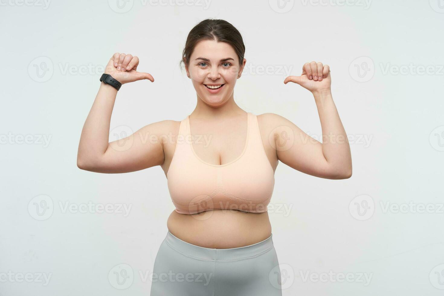 Studio shot of young happy cute dark haired fatty lady with ponytail hairstyle thumbing on herself while looking cheerfully at camera, standing over white background photo