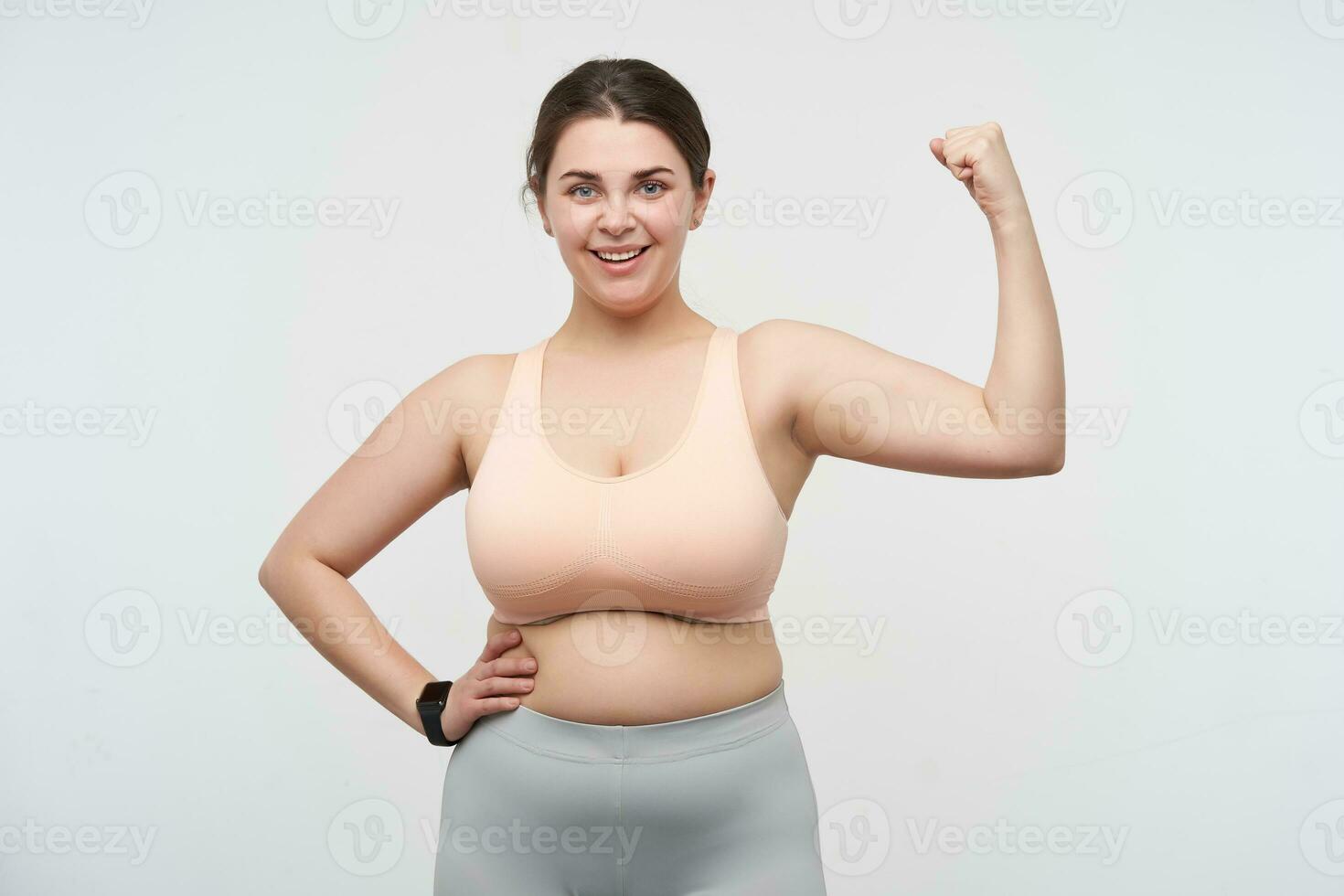 Indoor photo of young lovely brunette plump woman in sporty clothes looking gladly at camera while showing her biceps, standing against white background