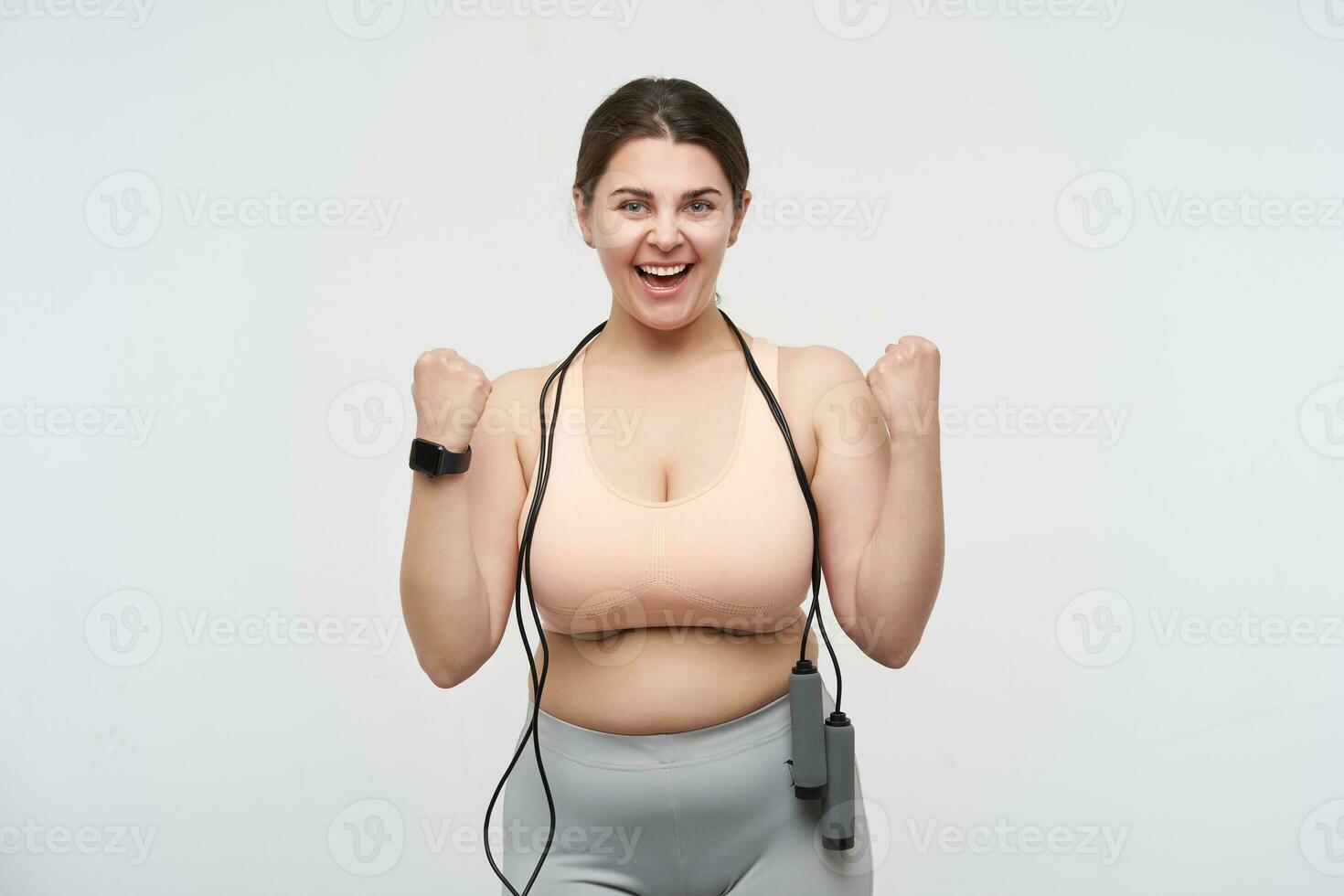 Indoor shot of cheerful young dark haired oversized woman with skipping rope raising happily her fist while rejoicing about nice working out, isolated over white background photo