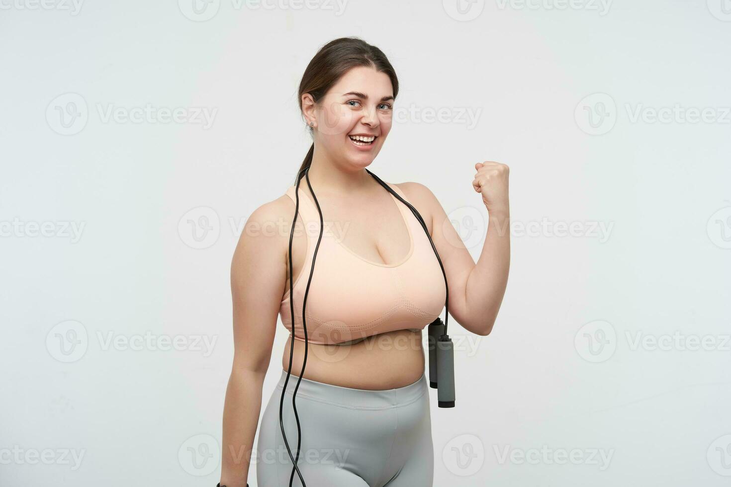 Cheerful young pretty brunette oversized lady in sporty wear looking joyfully at camera and raising hand in yes gesture, posing over white background. Concept of weight loss photo