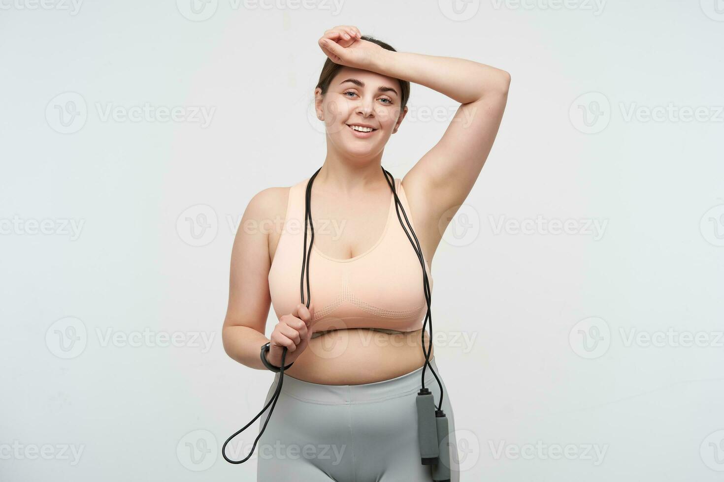 Positive young dark haired oversized woman dressed in sporty top and leggins wiping sweat of her forehead and smiling pleasantly while standing over white background with jumping-rope photo