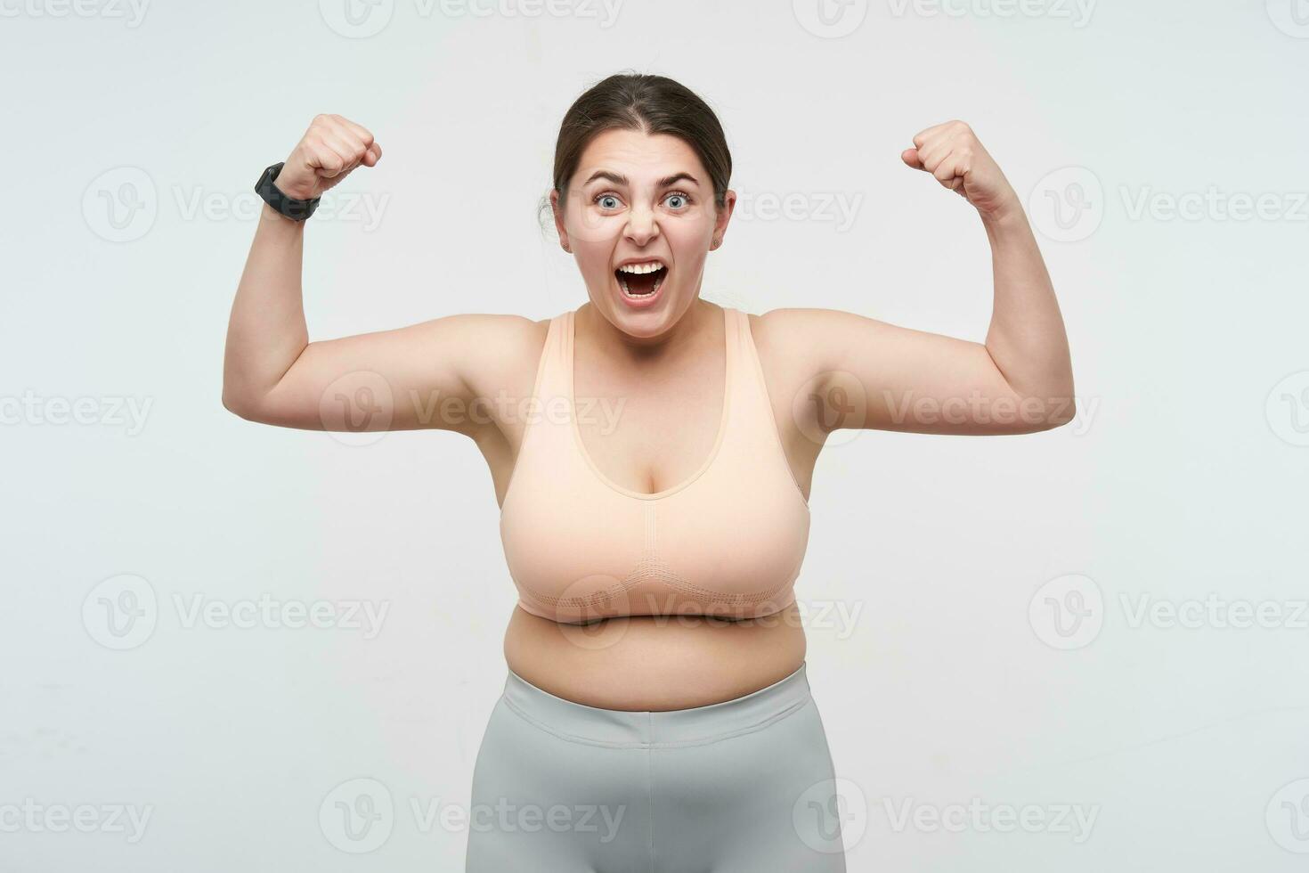 Expressive young brunette oversized female dressed in sporty clothes showing her power and looking excitedly at camera, posing over white background. Concept of weight loss photo