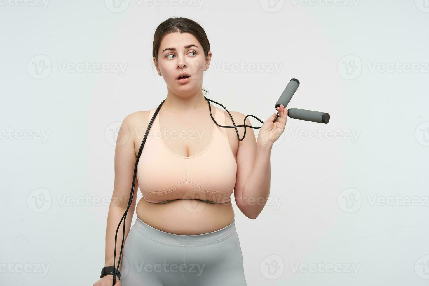 Indoor photo of thoughtful young dark haired oversized woman doing fitness and wearing jumping-rope on her neck while standing over white background. Body positive concept