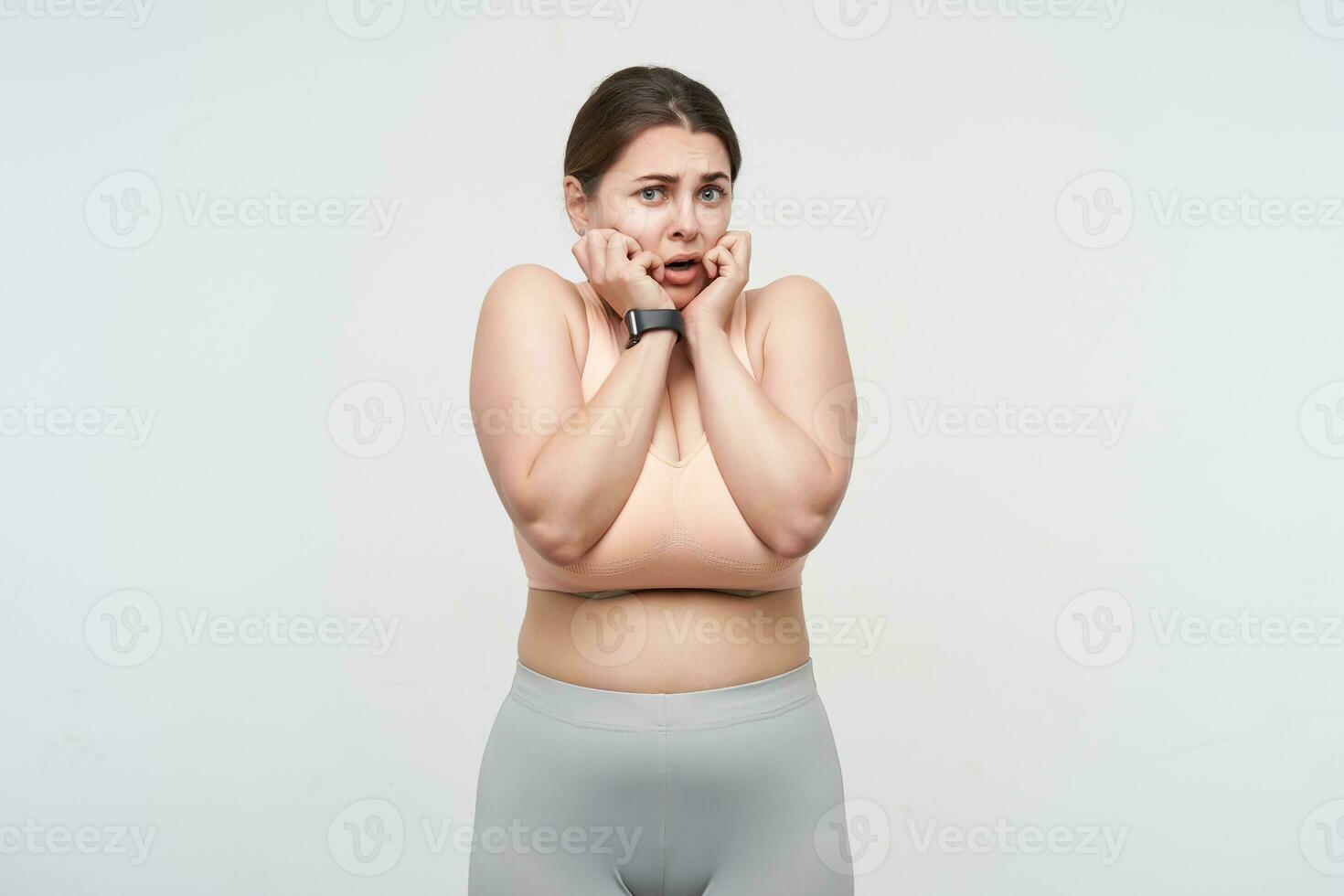 Bewildered young pretty brunette chubby lady dressed in sports bra and leggins keeping raised hands on her face while looking confusedly at camera. Concept of weight loss photo