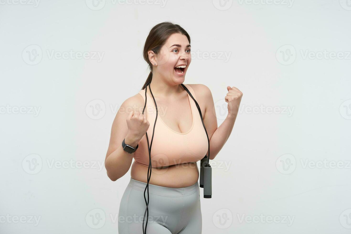 Side view of young overjoyed brunette fatty woman making sports and rejoicing about lost weight and raising happily hands, standing over white background with jumping-rope photo