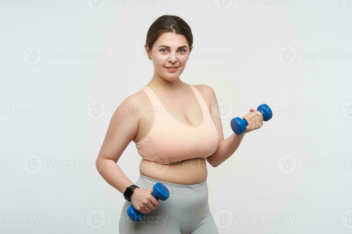 Studio photo of young positive oversized female exercising with dumbbells and looking gladly at camera while posing over white background. Concept of weight loss