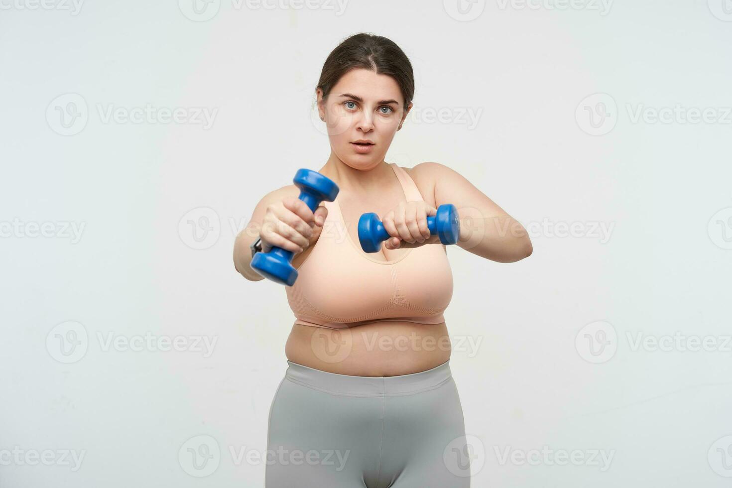 Studio shot of young plump oversized woman with ponytail hairstyle keeping dumbbells in raised hands while making fitness at home, isolated over white background photo