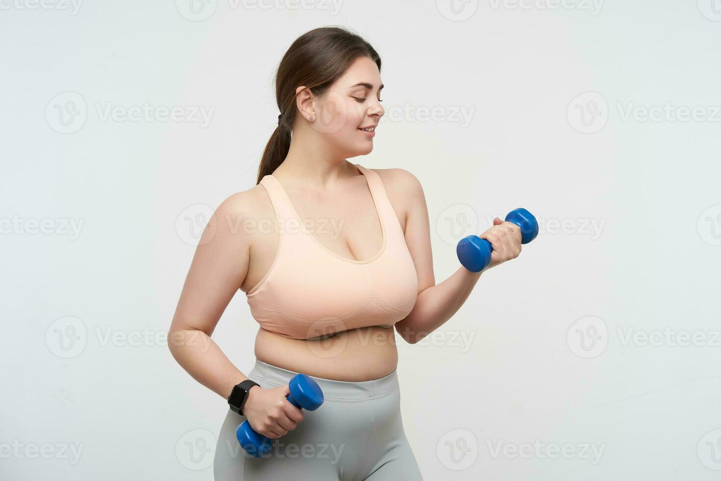 Indoor photo of young dark haired chubby woman dressed in sporty wear training her arms using little dumbbells while standing over white background. Body positive concept
