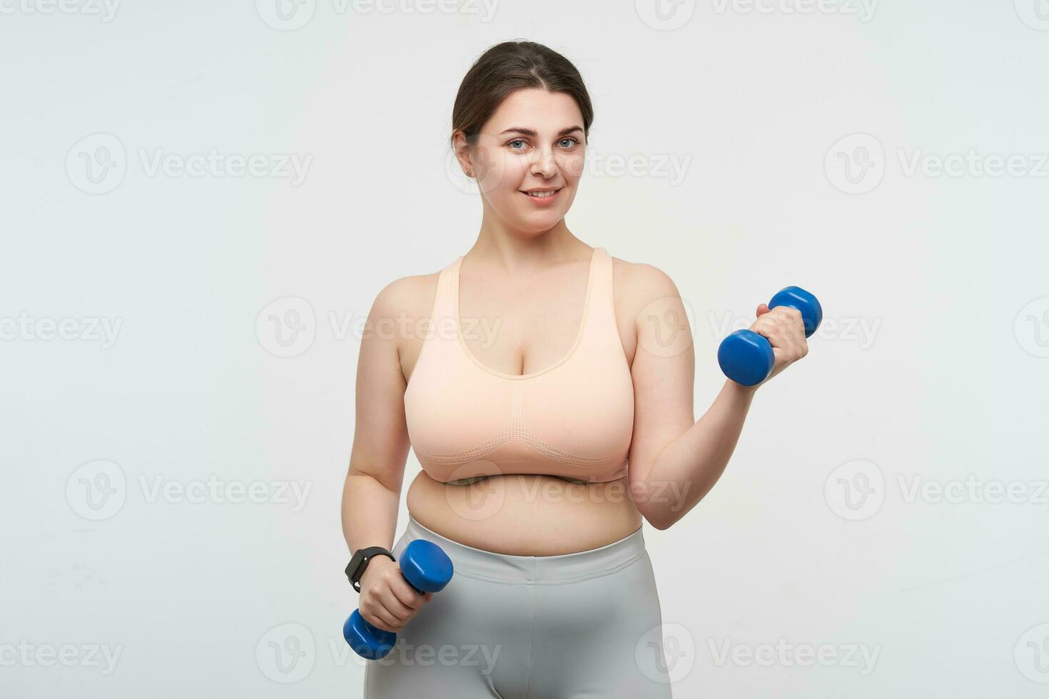 Cute young brunette chubby plump lady dressed in sporty clothes working out her hands with dumbbells and looking positively at camera while standing over white background photo