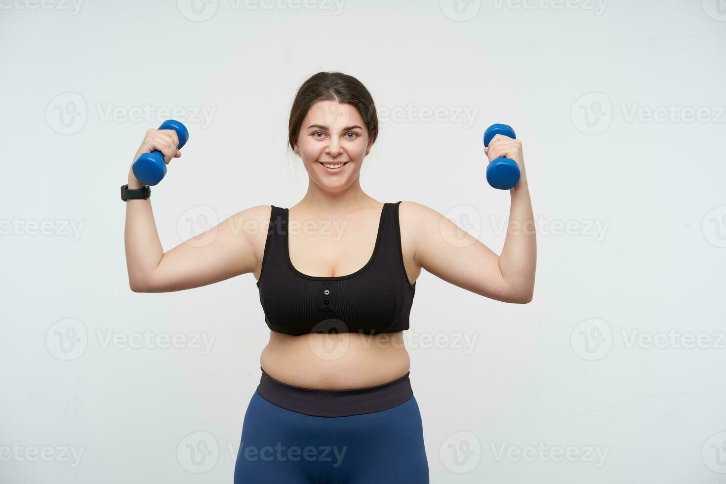 Happy young cute brunette cheerful with toothy smile plump woman lifting blue dumbbells and looking positively at camera, isolated over white background. Concept of self-made determination will-power photo