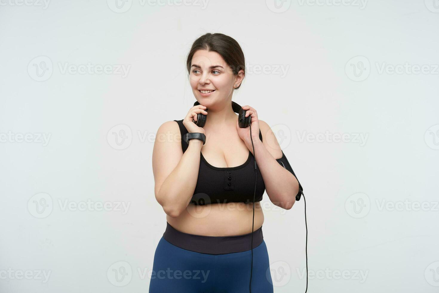 Pleased young cute brunette chubby lady with ponytail hairstyle holding raised hands on her earphones and looking positively aside, posing over white background photo
