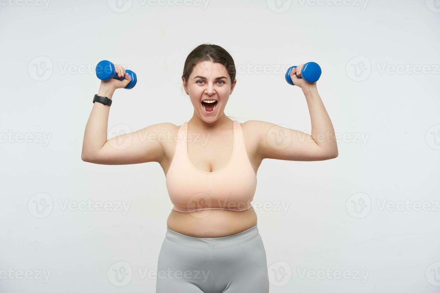 emocionado joven marrón peludo gordito hembra vestido en deportivo parte superior y leggins levantamiento emocionalmente manos con pesas mientras posando terminado blanco antecedentes. concepto de salir adelante por sí mismo determinación fuerza de voluntad foto