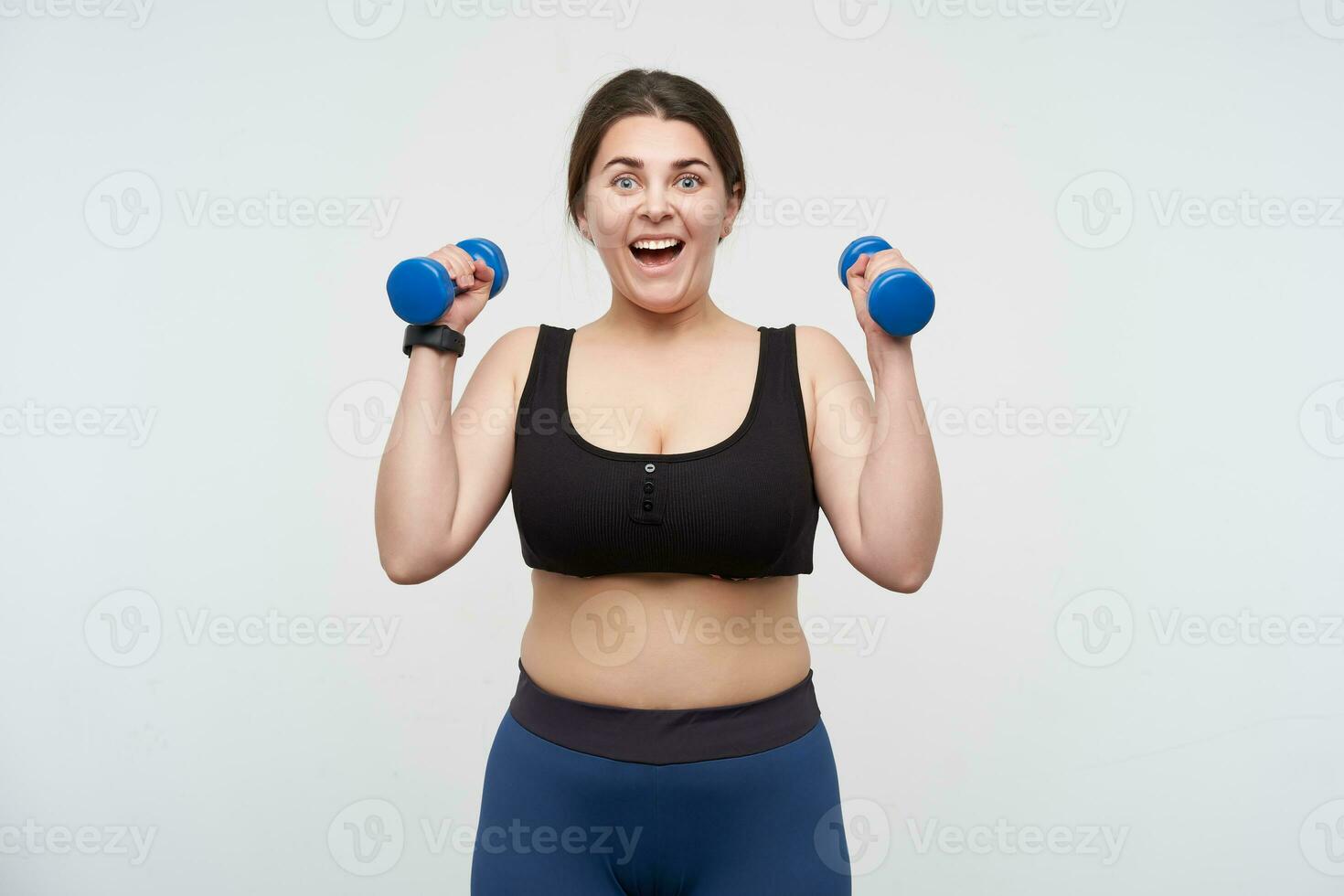 Agitated young cheerful oversized woman with casual hairstyle looking emotionally at camera while raising hands with blue dumbbells, posing over white background. Body positive concept photo