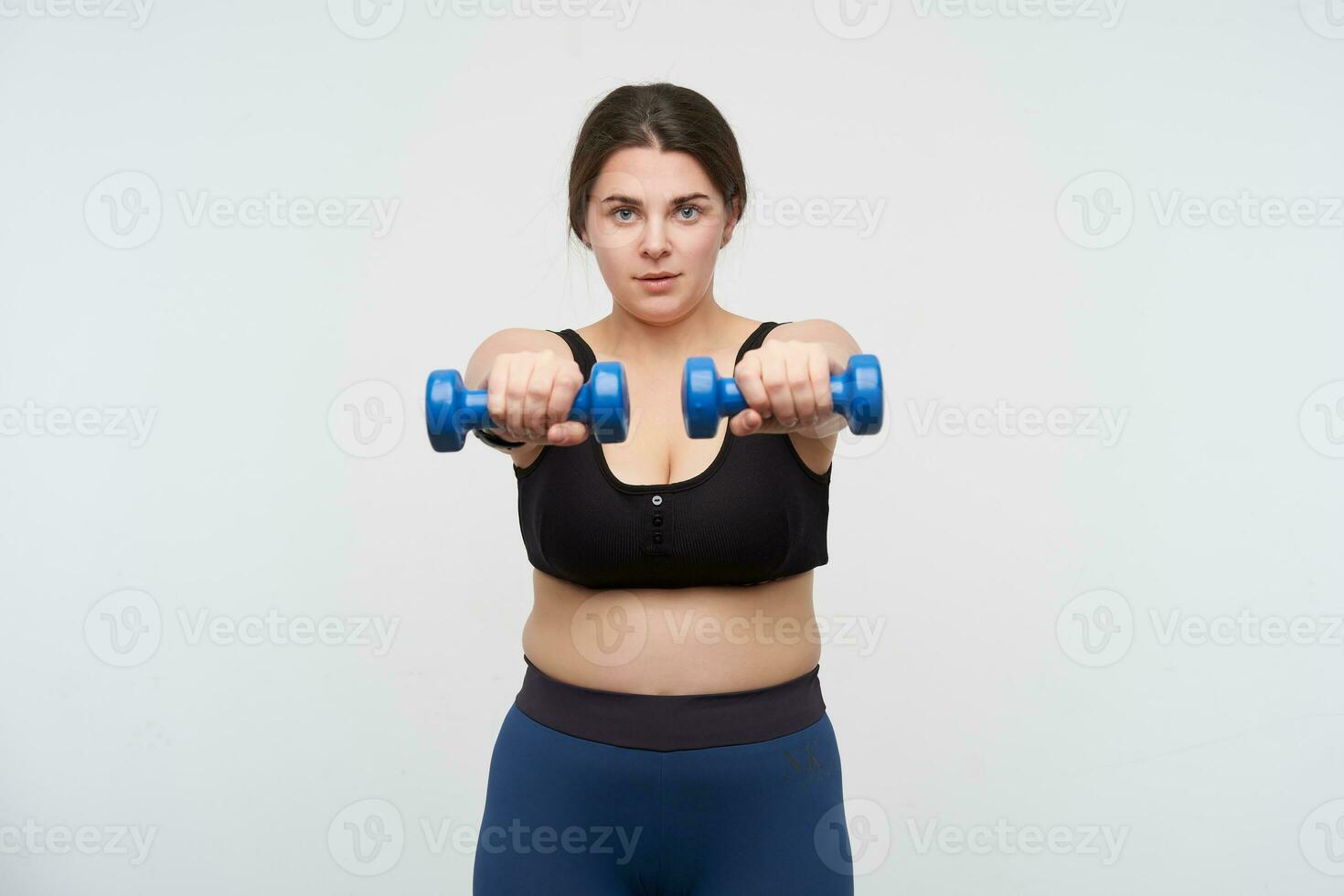 Concentrated young brunette chubby lady in sports bra and leggins keeping hands in front of herself while making exercises for hands, posing over white background photo