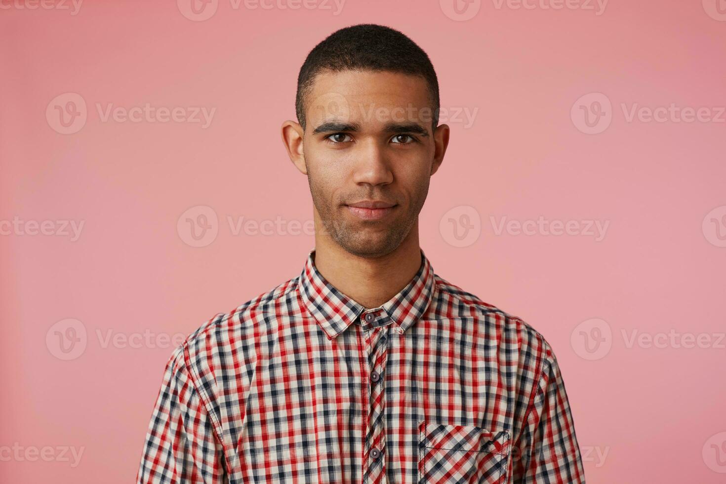 Close up of young attractive dark skinned guy in checkered shirt, looks at the camera with calm expression, stands over pink background. photo