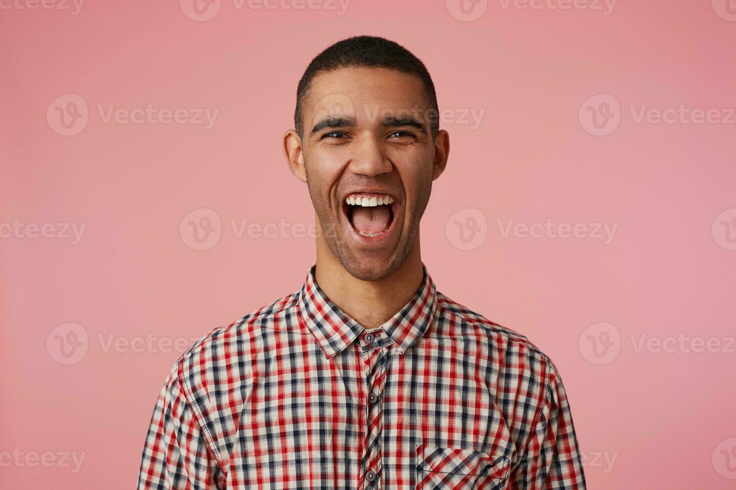 Portrait of laughing attractive dark skinned guy in checkered shirt, looks at the camera with happy expression and wide open mouth, hears funny joke, stands over pink background. photo